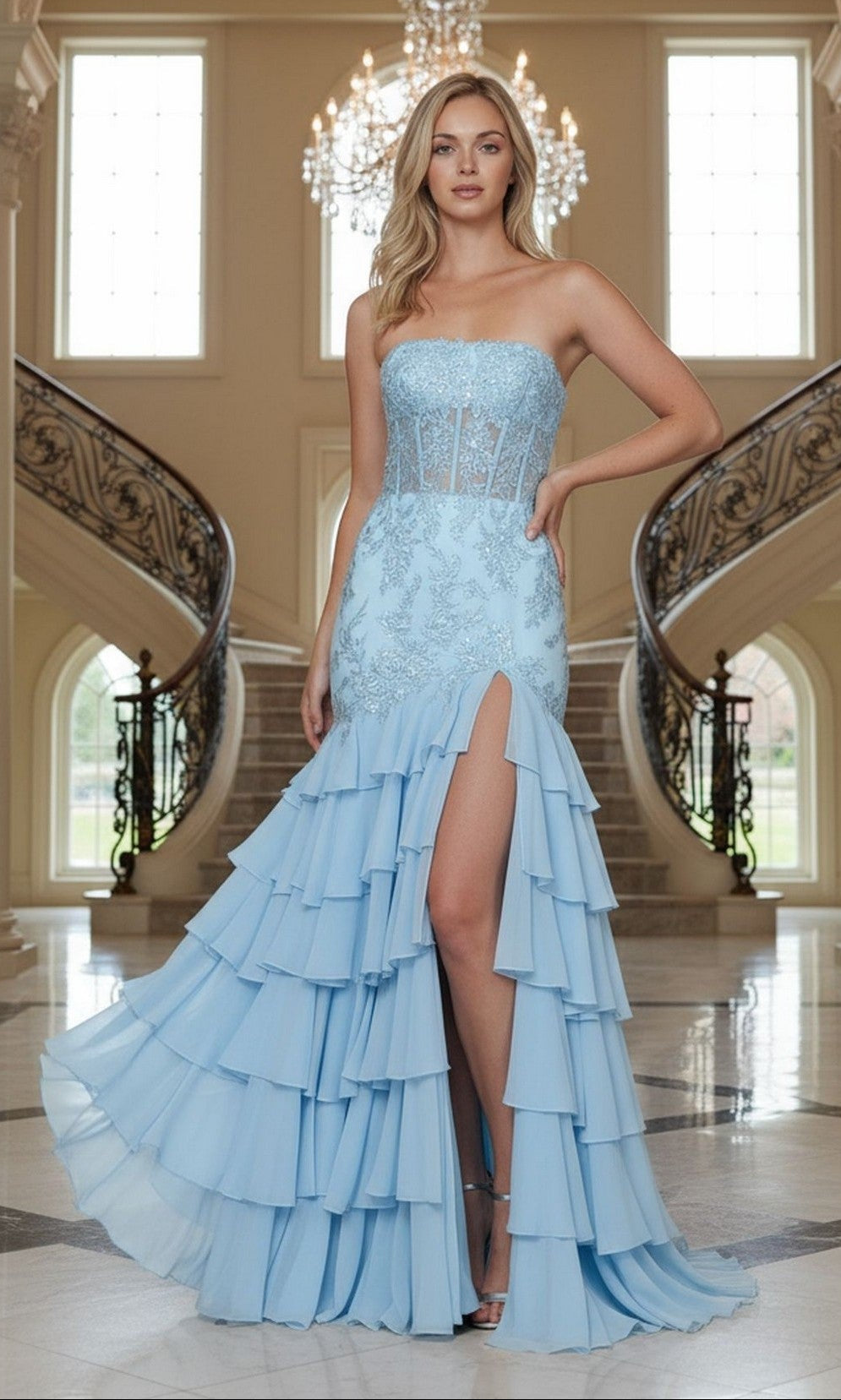 A woman stands in an elegant foyer wearing the blue Poly USA 25066 long prom dress, featuring a strapless lace-embroidered corset bodice, floral details, and a tiered ruffled chiffon skirt with a thigh-high slit.